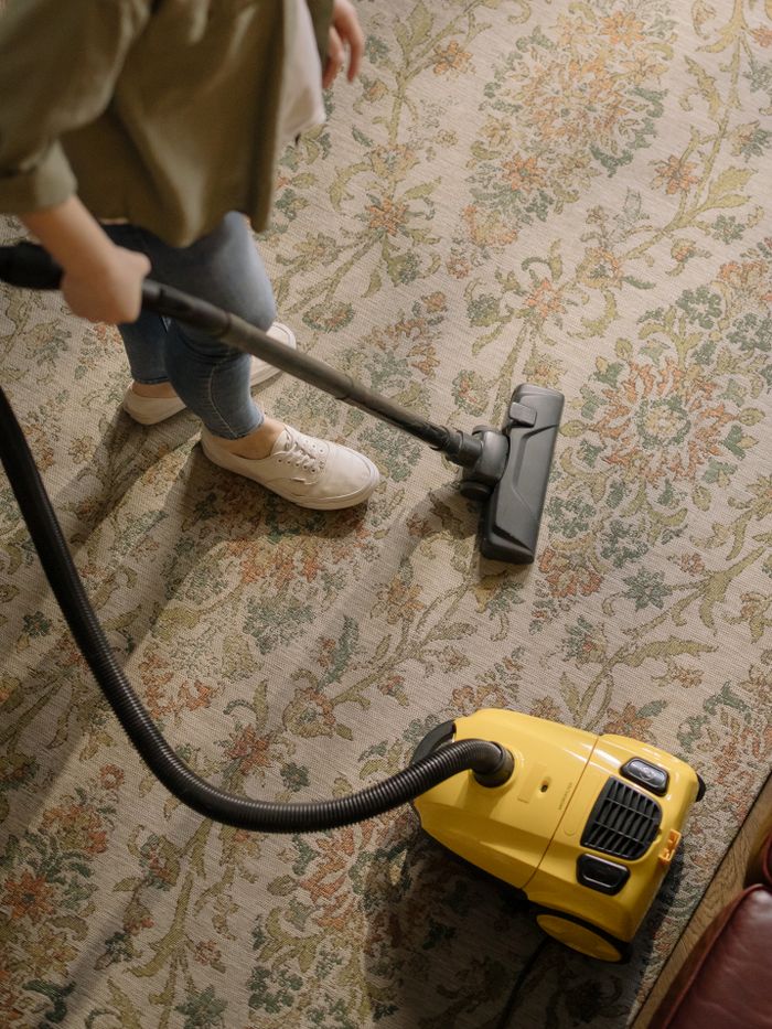 home-services-01 Personne en train de nettoyer un tapis fleuri avec un aspirateur jaune à l'intérieur.